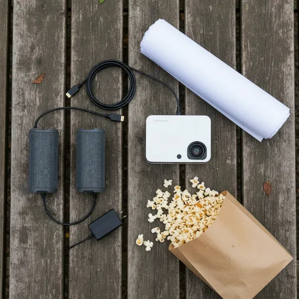 A collection of essential components for a DIY backyard theater, including a projector, a roll of white fabric, and portable speakers.